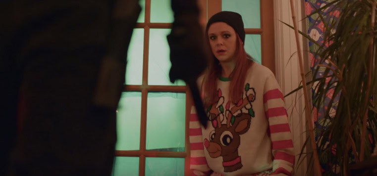 A person wearing a reindeer sweater and black beanie stands in a room with a green-glass door, exuding an unconventional surprise. A blurred figure in the foreground appears to wield a long object. In the background, decorated walls and a plant suggest an eerie holiday horror setting.