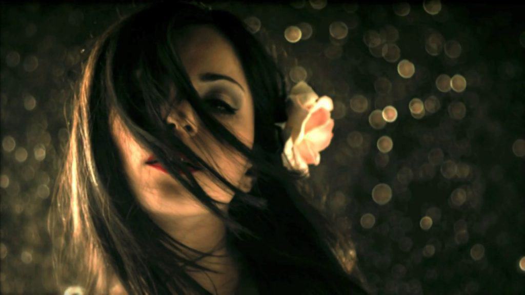 A woman with long dark hair, partially covering her face, poses against a sparkling, bokeh background. With a white flower in her hair and red lipstick, she exudes an unconventional allure, reminiscent of an enigmatic character from short films.
