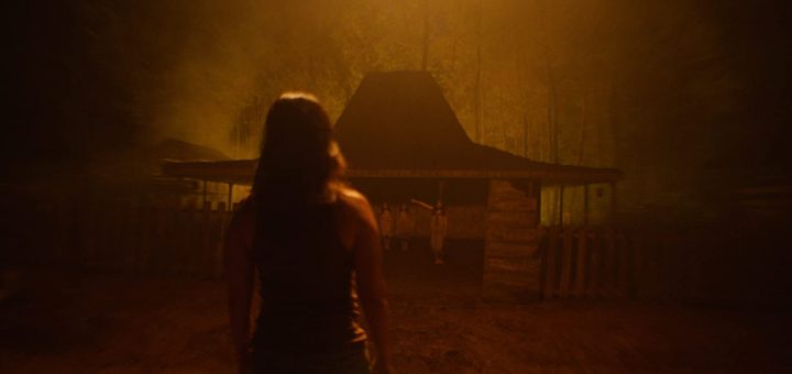 A person with long hair stands in the foreground at night, facing a dimly lit, rustic wooden house surrounded by trees and mist, with two figures visible near the entrance. The scene has an eerie, orange glow.