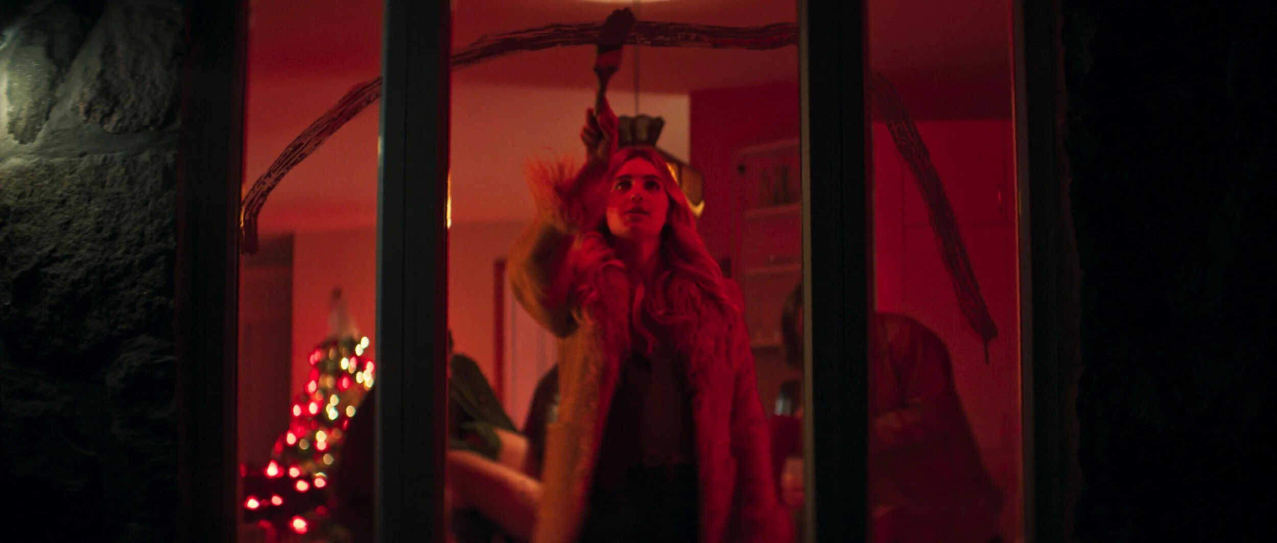 A person with long hair wearing a fur coat stands inside a warmly lit room, reaching up toward a branch hanging from the ceiling. In the background, a decorated Christmas tree glows amid festive, reddish lighting—evoking scenes from a classic Christmas movie.