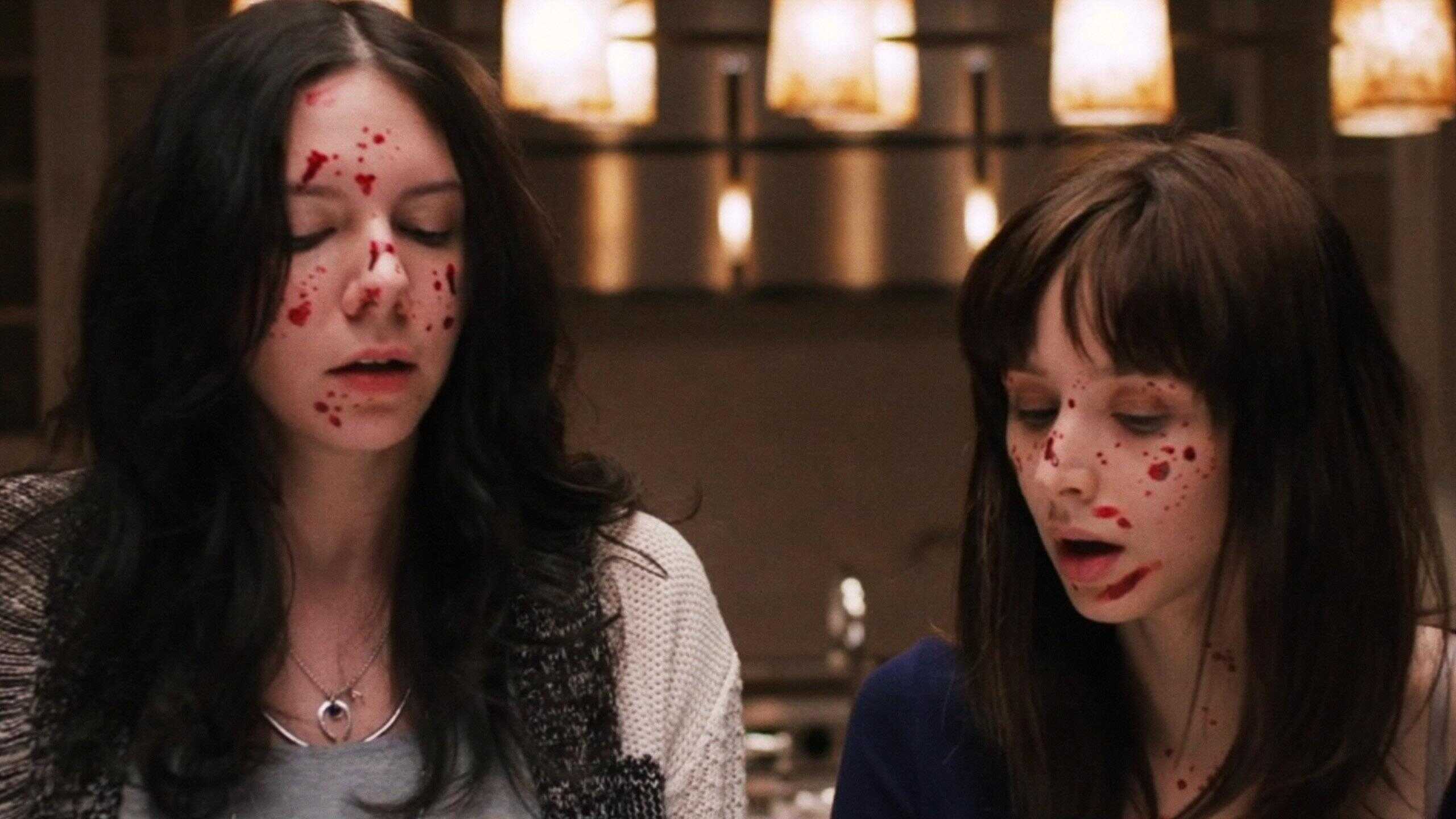 Two women with long dark hair and pale skin stand indoors with blood splattered on their faces. Both have their eyes closed or looking down, appearing distressed. Warm pendant lights hang in the blurred background.