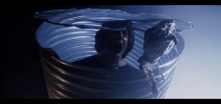 A person emerges from a large metal barrel with a mesh cover, holding up what appears to be a severed, bloody arm in a dark, eerie setting with blue lighting.