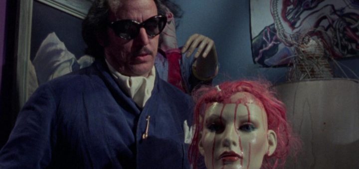 A man in dark sunglasses and a blue suit stands behind a mannequin head with red hair and fake blood running down its face, in a dimly lit, cluttered room with eclectic art and mannequin parts.