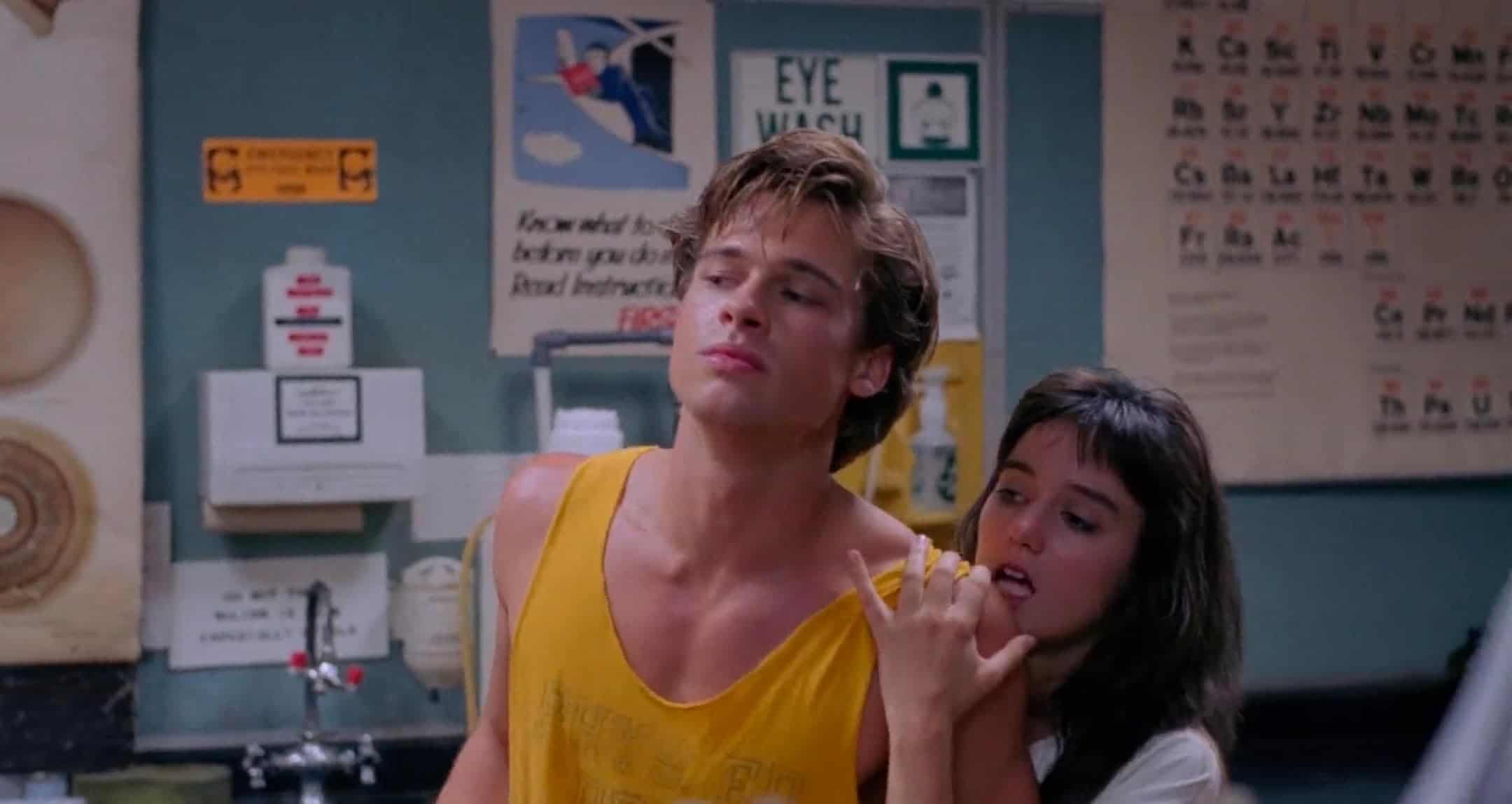 A young man in a yellow tank top stands with a relaxed expression as a young woman leans on his shoulder, holding his arm. Behind them are wall posters, safety signs, and an eye wash station in a classroom or lab setting.