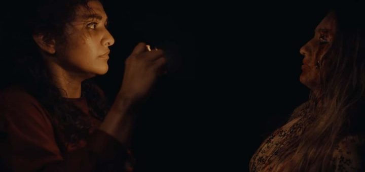 Two women face each other in a dark setting; one holds a lighter up, illuminating both their faces. The other woman has long hair and blood on her face, creating a tense atmosphere.