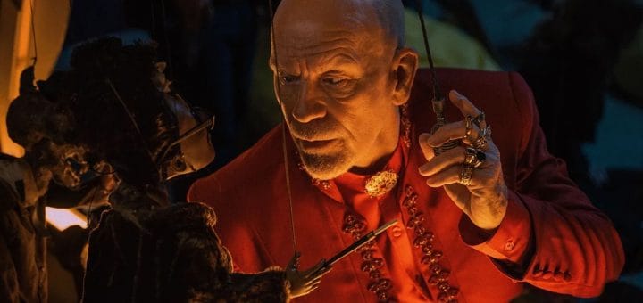 A bald man in a red suit with ornate rings and a beaded necklace intently manipulates a puppet, holding its strings in one hand and looking closely at its face in a dimly lit setting.
