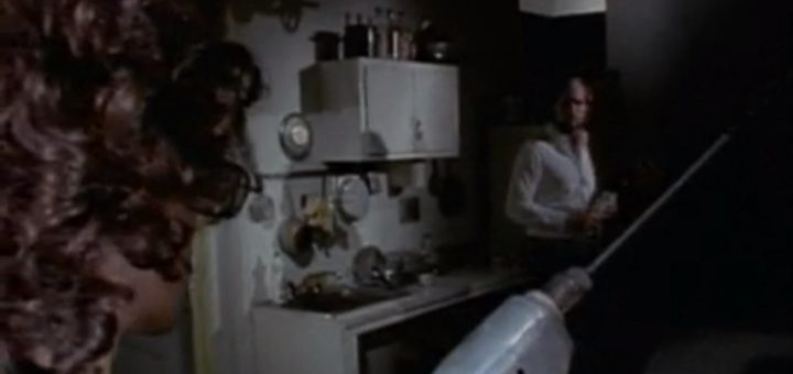 A person with curly hair holds a large electric drill in the foreground, aiming at another person standing in a dimly lit kitchen area cluttered with dishes and bottles.