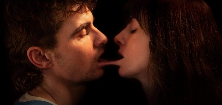 A close-up of a man and a woman facing each other against a dark background. The woman’s tongue is unusually elongated, stretching into the man’s mouth. Both have their eyes closed, suggesting intimacy.