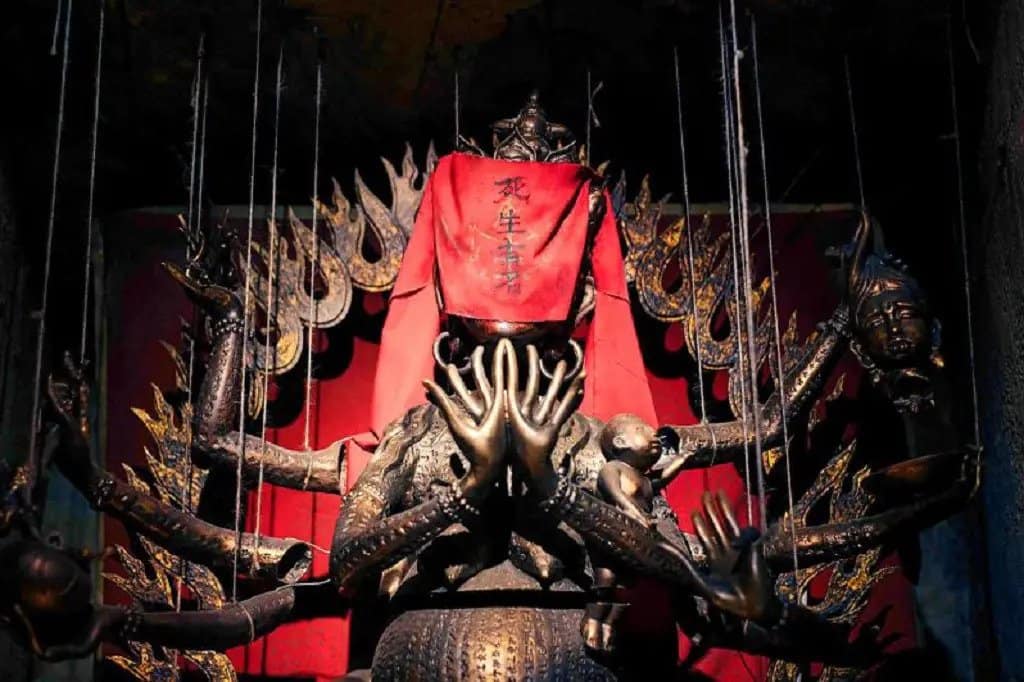 A multi-armed statue with a red cloth covering its face, surrounded by intricate golden details and additional heads, stands against a red background. The hands are raised in various gestures.