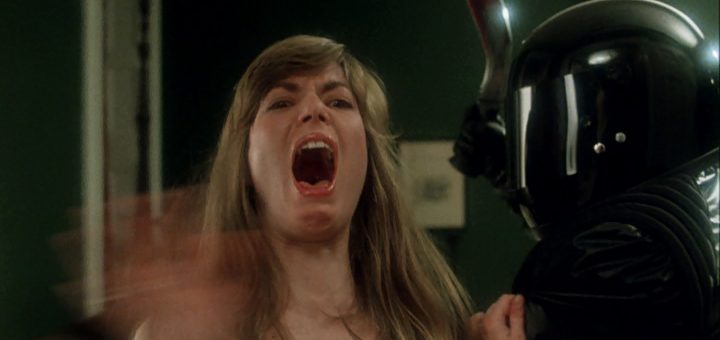 A woman with long hair screams in fear with her mouth wide open, while a person in black armor and a shiny helmet stands beside her, grabbing her arm. The scene appears intense and dramatic.