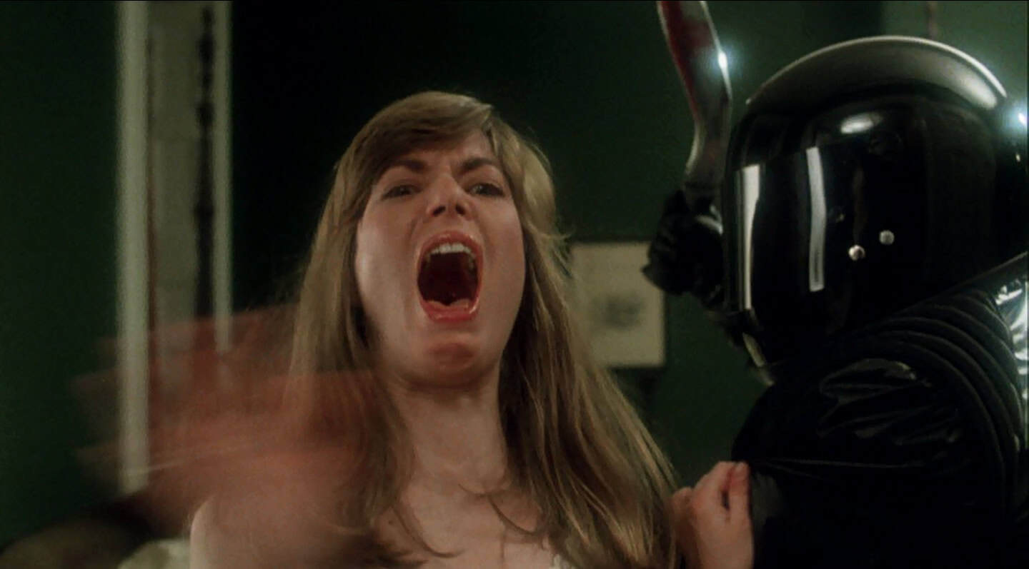 A woman with long hair screams in fear with her mouth wide open, while a person in black armor and a shiny helmet stands beside her, grabbing her arm. The scene appears intense and dramatic.