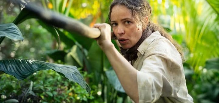 A woman in a light shirt crouches in dense green jungle foliage, clutching a long spear with a determined expression—ready for an adventurous birthday celebration as sunlight filters through the leaves.