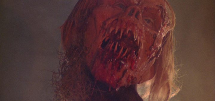 A gruesome, partially decomposed face with long blond hair, exposed bone, and blood-covered mouth forms a horrifying scene—perfect for a twisted Happy Birthday scare in dim lighting.