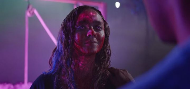 A woman with long hair stands in colorful, moody lighting reminiscent of Color Out Of Space, her face and neck covered in red markings or wounds. She looks ahead with a serious expression, framed by a blurred background of purple and blue tones.