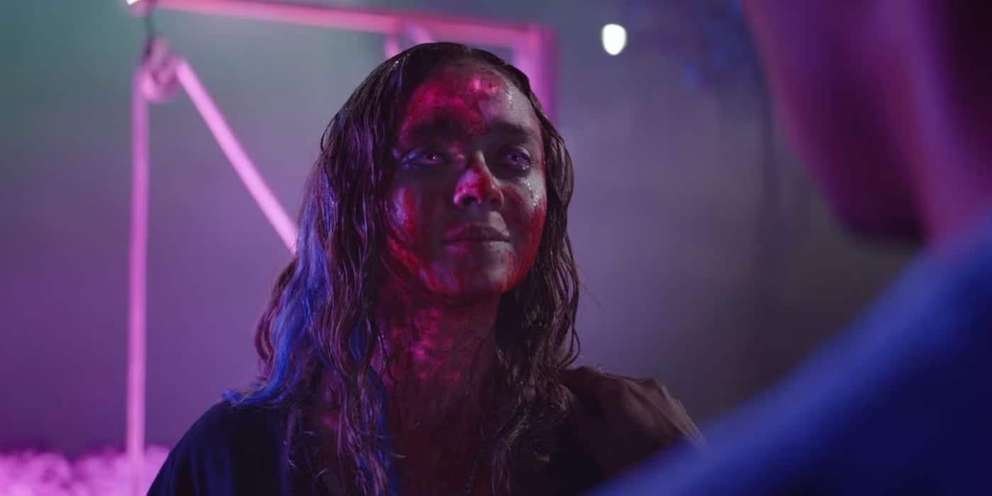 A woman with long hair stands in colorful, moody lighting reminiscent of Color Out Of Space, her face and neck covered in red markings or wounds. She looks ahead with a serious expression, framed by a blurred background of purple and blue tones.