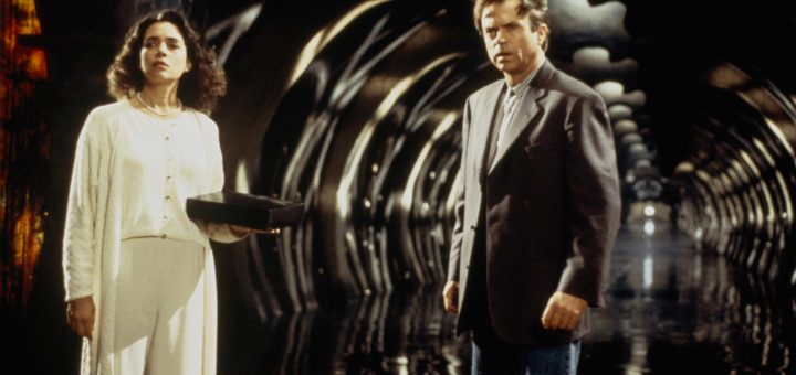 A woman in white holds a box while standing next to a man in a suit inside a dark, tunnel-like space with reflective floors and curved, illuminated arches, evoking the eerie atmosphere of A Nightmare On Elm Street 4: The Dream Master.