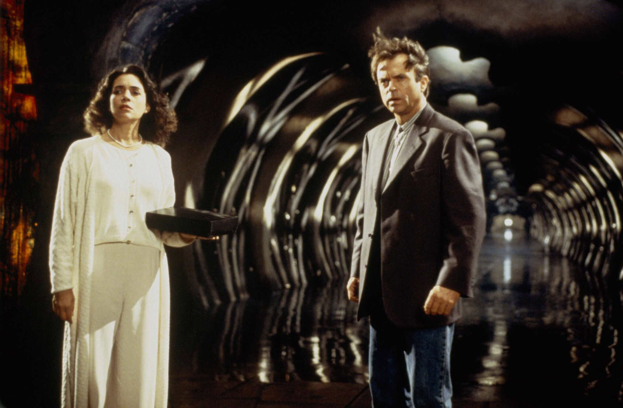 A woman in white holds a box while standing next to a man in a suit inside a dark, tunnel-like space with reflective floors and curved, illuminated arches, evoking the eerie atmosphere of A Nightmare On Elm Street 4: The Dream Master.