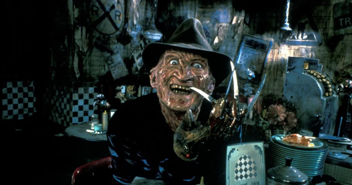 A scarred man with a brown hat and bladed glove grins menacingly in a dim, cluttered room, holding a cigarette between his metal claws—a chilling figure straight out of A Nightmare On Elm Street 4: The Dream Master.