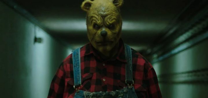 A person wearing a bear mask, red and black plaid shirt, and denim overalls stands in a dimly lit hallway, their menacing expression evoking the unsettling atmosphere of Color Out Of Space.