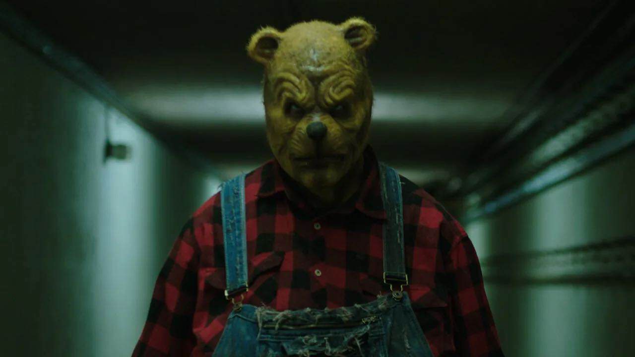 A person wearing a bear mask, red and black plaid shirt, and denim overalls stands in a dimly lit hallway, their menacing expression evoking the unsettling atmosphere of Color Out Of Space.