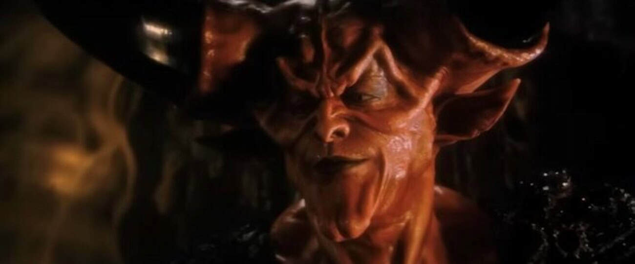 A close-up of a red-skinned, horned demon with sharp facial features and a sinister expression, wearing dark armor, in a dimly lit setting.