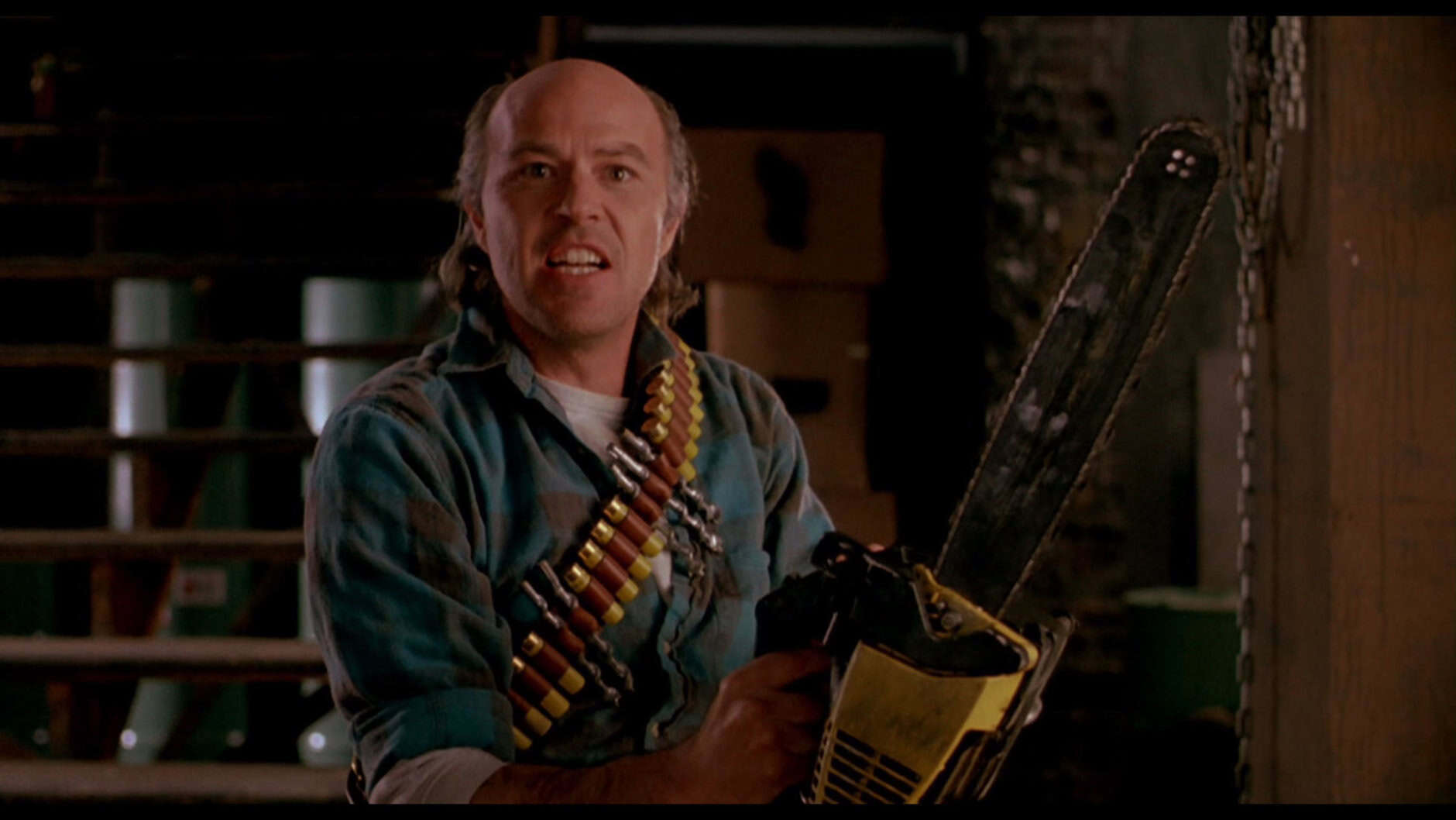 A man in a plaid shirt with a shotgun shell bandolier holds a large chainsaw indoors, looking intensely forward. Cardboard boxes and stairs are visible in the background.