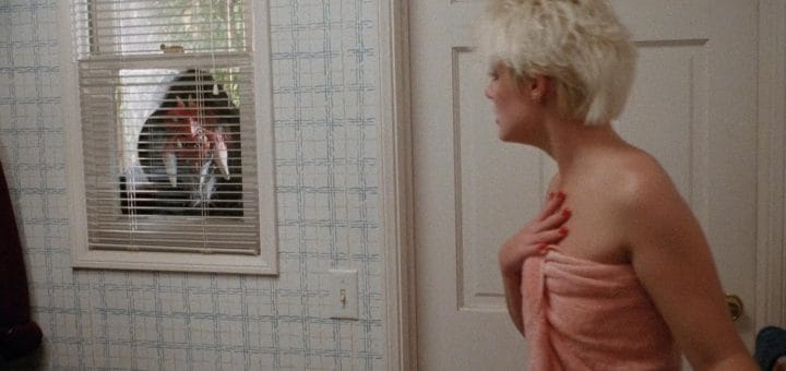 A woman in a towel looks startled as she sees a hooded figure staring at her through a window with blinds in a bathroom, evoking the chilling suspense of a scene straight out of the 1988 horror movie Hack-O-Lantern.