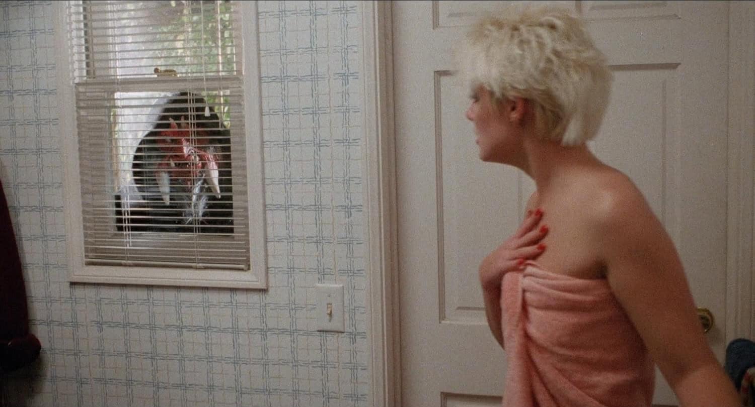 A woman in a towel looks startled as she sees a hooded figure staring at her through a window with blinds in a bathroom, evoking the chilling suspense of a scene straight out of the 1988 horror movie Hack-O-Lantern.