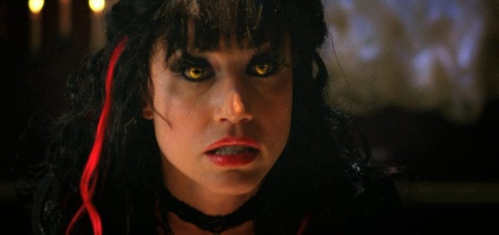 A woman with yellow cat-like eyes, red streaks in her black hair, red lipstick, and a black choker looks intensely forward, evoking the eerie atmosphere of a 2009 horror movie. The background is dimly lit with blurred lights, reminiscent of Night of the Demons.