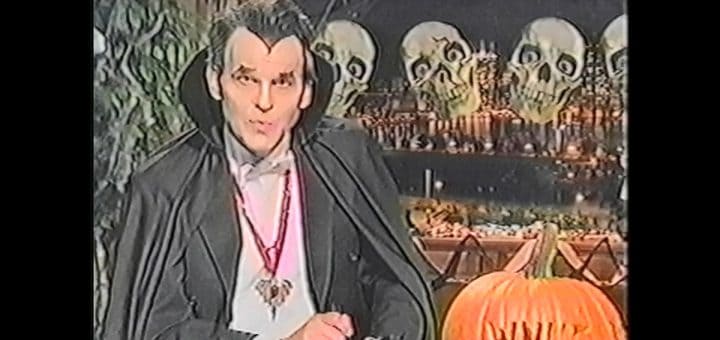 A person dressed as a vampire sits in front of Halloween decorations, including skulls, a Hack-O-Lantern, and a pumpkin carved with the letters “WNUF.”.