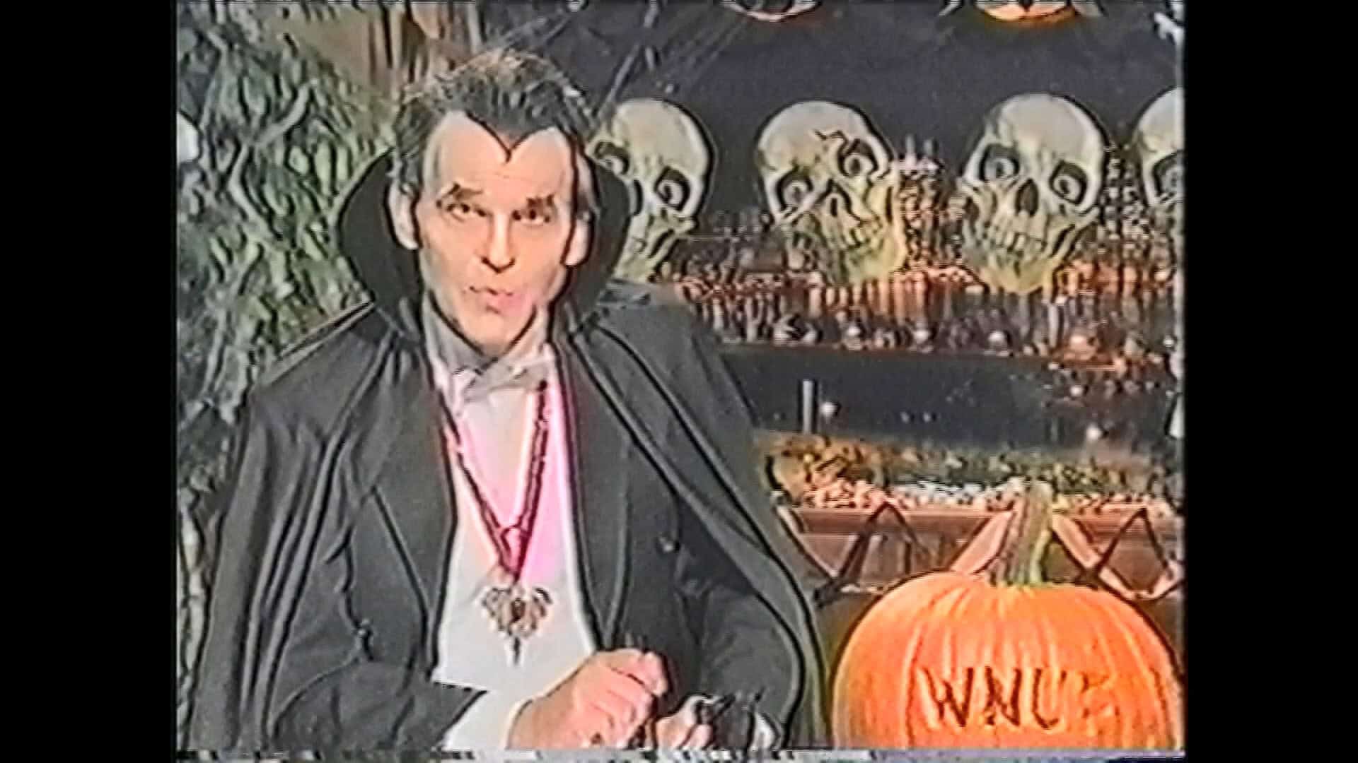 A person dressed as a vampire sits in front of Halloween decorations, including skulls, a Hack-O-Lantern, and a pumpkin carved with the letters “WNUF.”.
