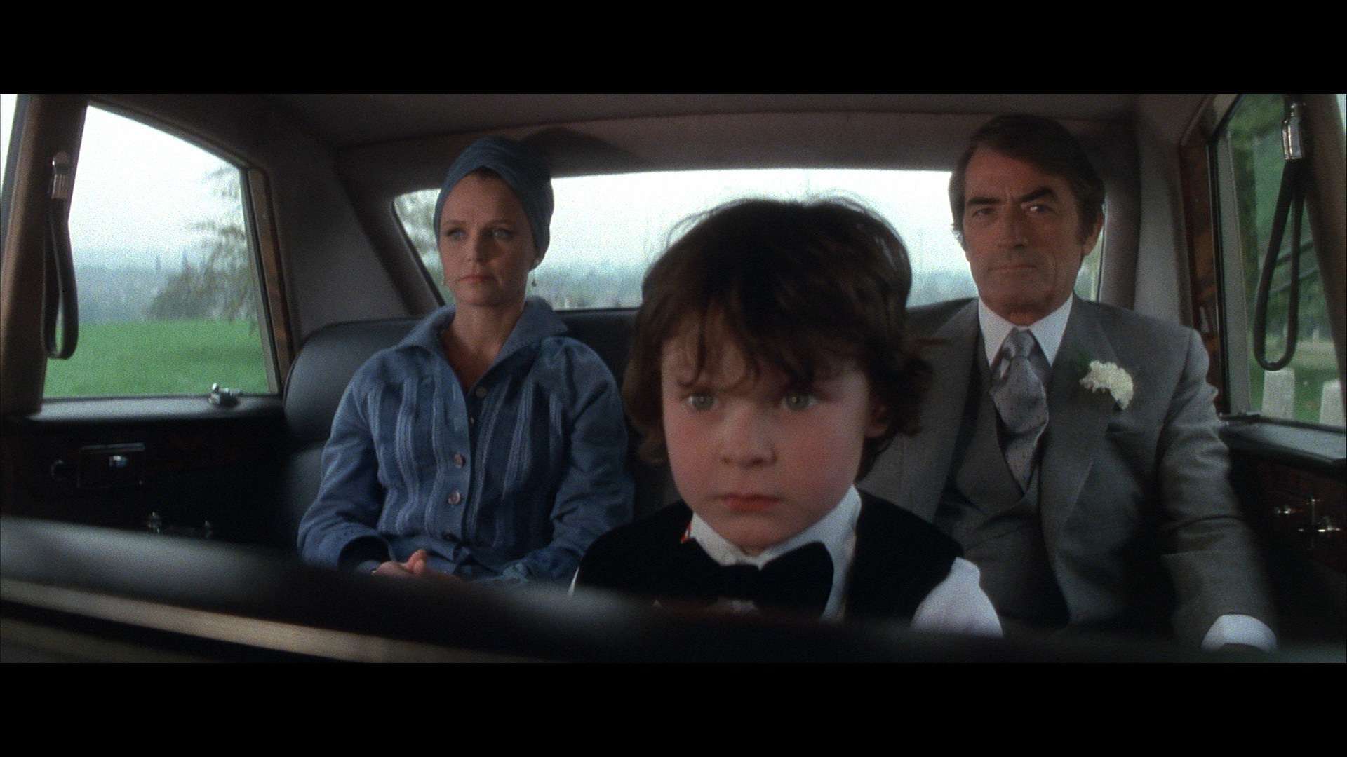 A young boy with short dark hair sits in the backseat of a car, flanked by a woman in a blue outfit and headscarf and a man in a suit with boutonniere. They all appear serious, as if focused on the next chilling episode of their favorite horror movie podcast playing softly in the background.