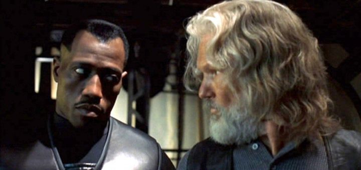 Two men stand side by side. The man on the left, with short hair and dark clothing, looks as sharp as a blade. Meanwhile, the man on the right, sporting long white hair and a beard under a dark vest over his shirt, glances thoughtfully at his companion.