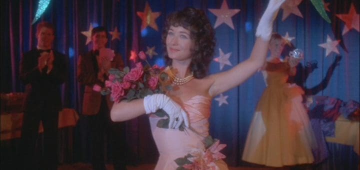 A woman in a strapless pink dress and white gloves holds a bouquet of pink flowers on a stage, reminiscent of Mary Lou's infamous Prom Night 2. She waves to an audience beneath blue curtains adorned with star decorations, as people stand behind her, applauding with an eerie enthusiasm.