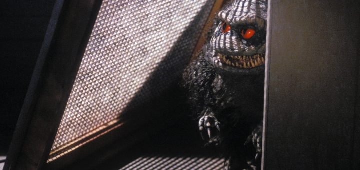 A small, menacing creature with red eyes and sharp teeth stands in a dimly lit space, partially obscured by a vent-like structure. Its dark, furry body contrasts with the light filtering through the perforated surface.