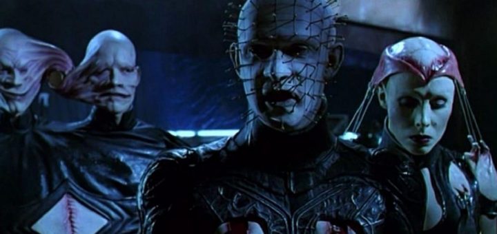 A group of eerie, otherworldly characters with distorted features stand in a dimly lit setting. The central figure has pins in its head, and others have elongated faces or unusual headgear, all wearing dark, detailed clothing. The mood is sinister and surreal.