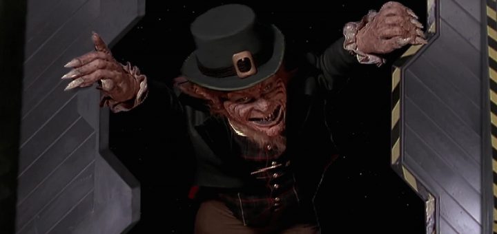 A menacing creature dressed as a leprechaun, with a green top hat and sharp teeth, floating in a dark space with outstretched arms. The background features sci-fi elements and an open hatch.