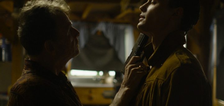 In a dimly lit room, echoing the tense atmosphere of a hospital massacre, two men stand face-to-face—one with a handgun aimed at the other's throat. The gunman's serious expression contrasts sharply with the other’s slight head tilt, set against wooden elements and a blurred window.