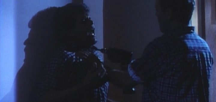 A dimly lit scene shows a tense confrontation between two people. One, partially in shadow, wields a drill like a carpenter gone rogue against the other's chest. Their expressions suggest fear and aggression, highlighting a dramatic moment reminiscent of woodworking gone awry.