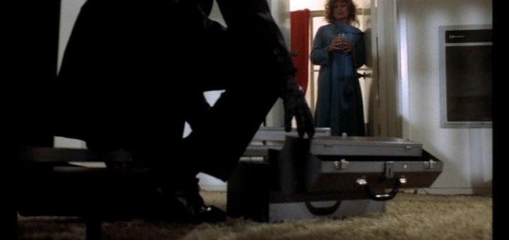 A person in a trench coat and gloves is crouching, opening a briefcase on the floor. In the background, a woman stands in a doorway, holding an object, looking towards the person. The room is dimly lit and carpeted.