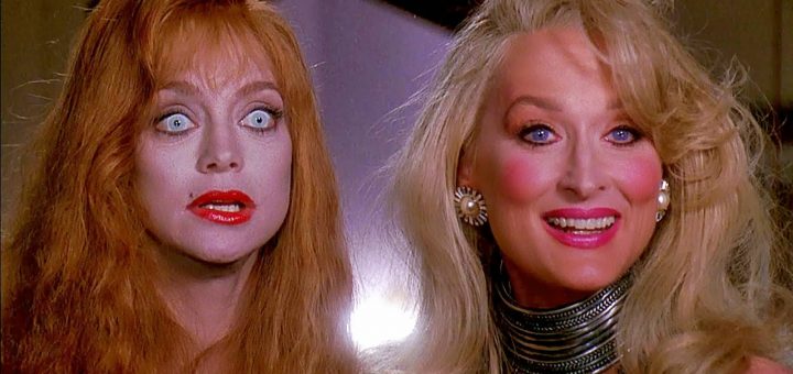 Two women stand side by side; one has pale skin, wide blue eyes, red lips, and red hair, appearing shocked. The other woman, with blonde hair and bright makeup, smiles confidently, wearing statement jewelry and a glamorous outfit.