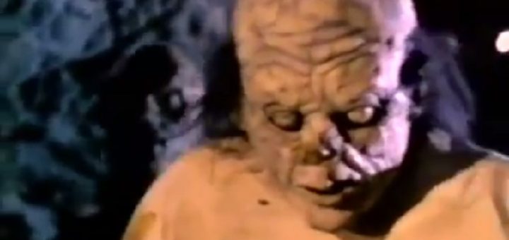 A person wearing a grotesque, lumpy, monster-like mask with visible veins and blemishes, dressed in a light-colored shirt, stands in a dimly lit setting.