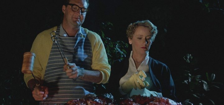 A man and woman stand by a grill at night. The man wears glasses, an apron, and holds barbecue tools, while the woman, in gloves, looks at the food with a serious expression. Grilled meat is piled in front of them.