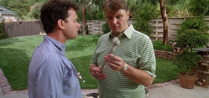 Two men stand in a backyard; one, in a striped green polo shirt, closely examines a bone, while the other, in a blue shirt, looks at him. Trees and a lawn are visible in the background.
