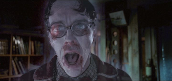 A pale, ghostly man with wet, slicked hair, wide eyes behind round glasses, and an open mouth appears shocked or frightened in a dimly lit room with shelves and machinery in the background.