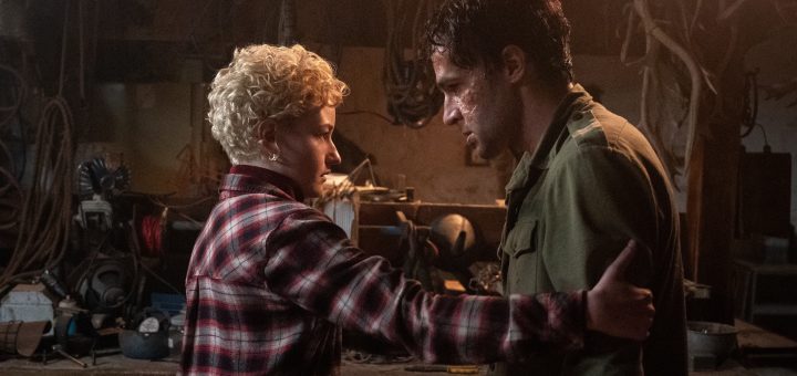 A woman with short curly blond hair and a red plaid shirt holds the arm of a man with disheveled hair and a bruised face in a dimly lit, cluttered workshop. They look intensely at each other.