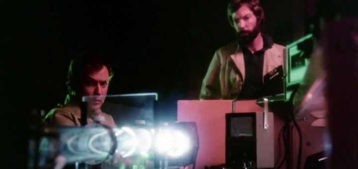 Two men in lab coats work with scientific equipment, illuminated by glowing green and blue lights in a dark laboratory setting—a scene reminiscent of Murder By Phone. One man sits, focused, while the other stands behind machinery.