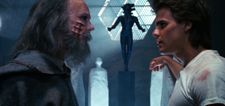 A scarred older man in a cloak faces a younger man in a white shirt in a dark room with statues and a geometric, horned figure on a pedestal in the background. They appear to be having an intense conversation.