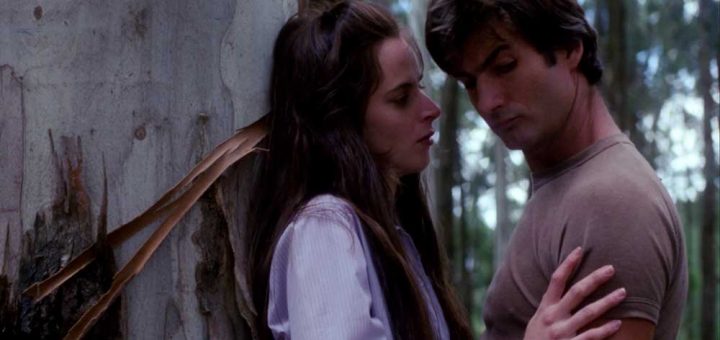 A woman and a man stand close to each other by a tree in a forest. The woman leans against the tree, gently touching the man's chest, while both look down with serious expressions.