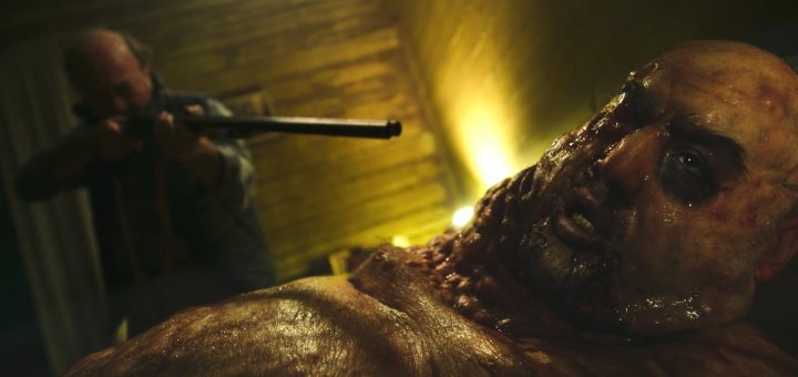 A man with severe, grotesque facial and body mutations lies in the foreground, looking distressed, while a second man in the background points a shotgun at him inside a dimly lit, wooden room.